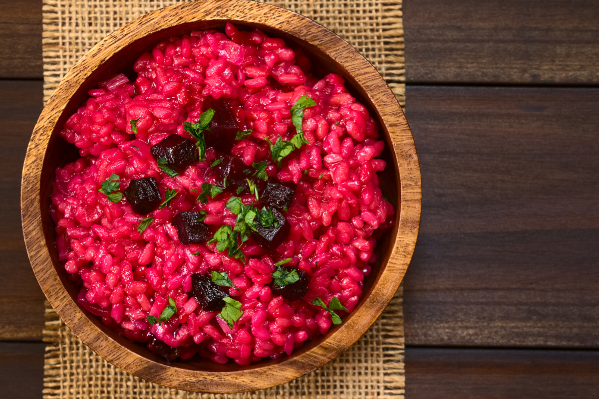 A wooden bowl filled with vibrant pink beet and barley risotto, garnished with chopped parsley and beet cubes, placed on a rustic woven mat.