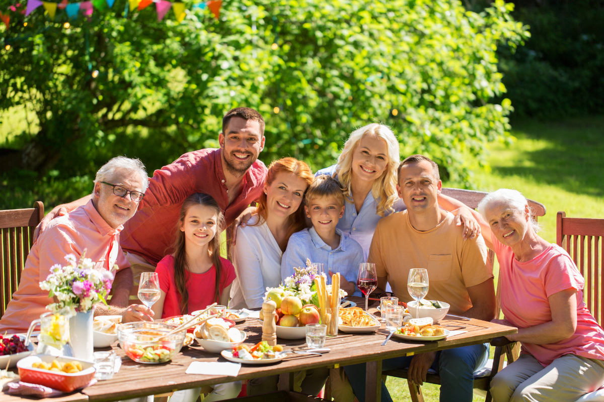 A multi-generational family smiling and enjoying an outdoor meal at a picnic table in a lush garden, with colorful bunting in the background.