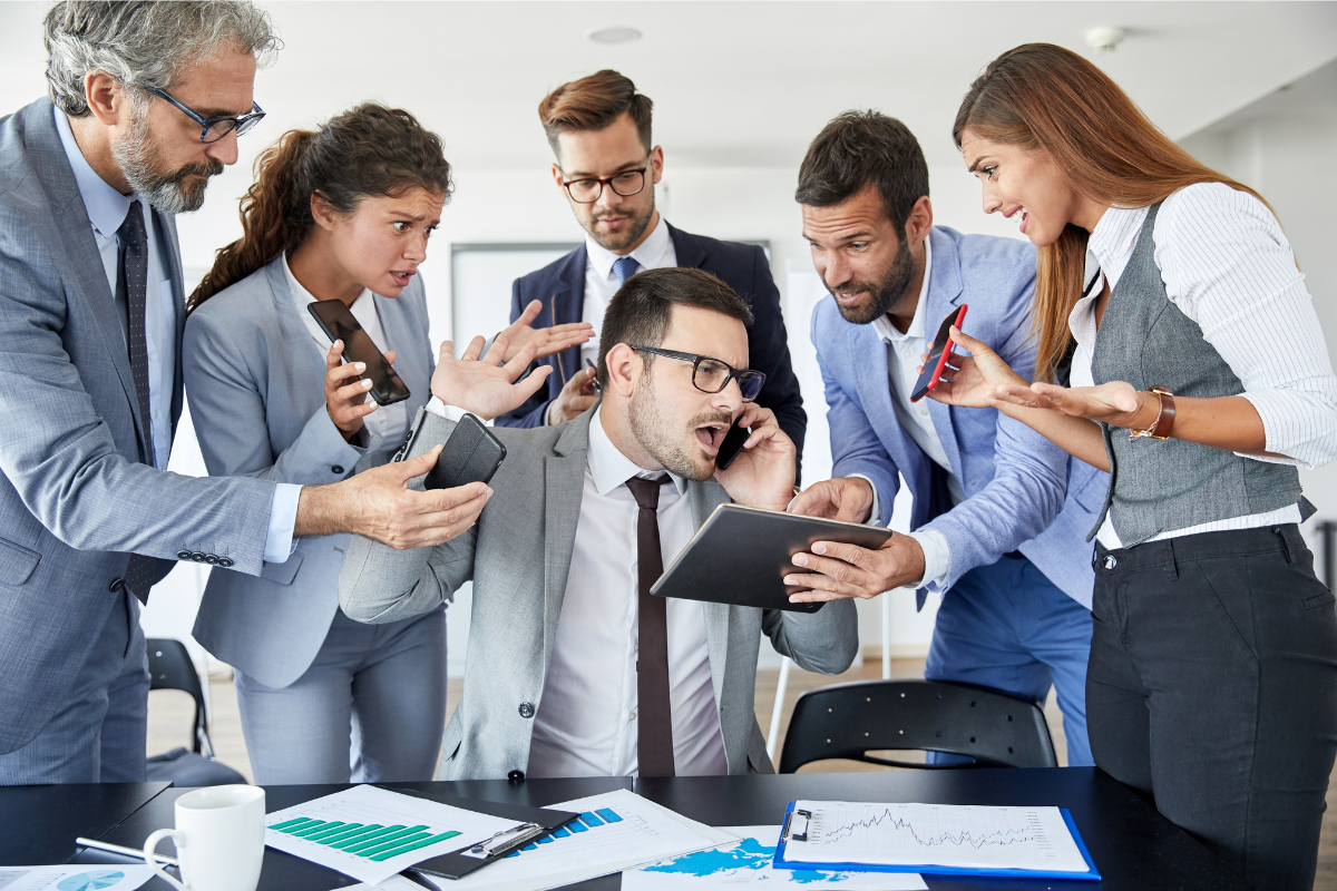 A stressed businessman surrounded by colleagues demanding his attention, holding multiple devices and documents in a busy office setting.