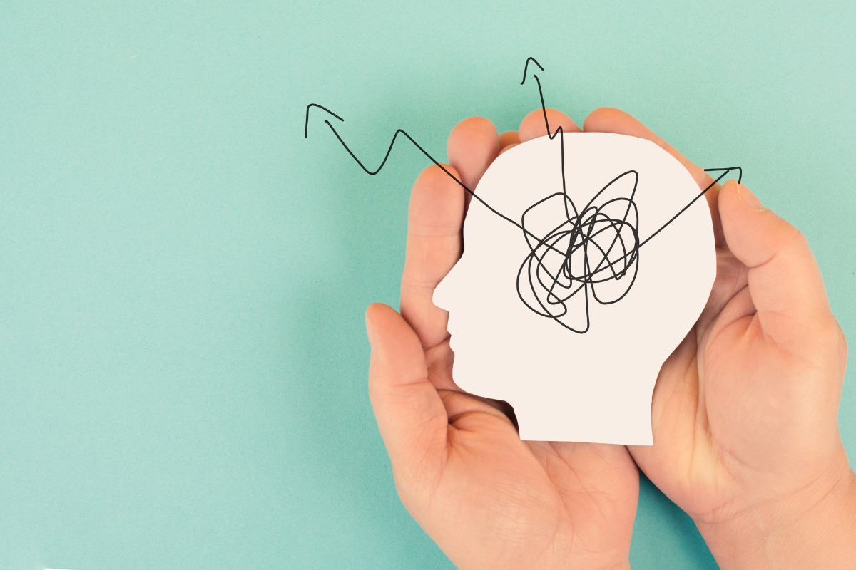 Hands holding a paper cutout of a head with scribbled lines inside, symbolizing mental confusion or stress, on a turquoise background with arrows pointing outward.