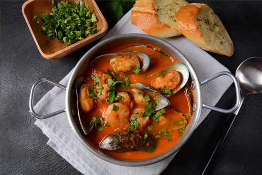 A grey casserole filled with seafood stew, featuring mussels, with slices of bread nearby.