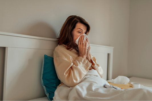 A woman sitting in bed, holding a tissue to her nose, appearing unwell.