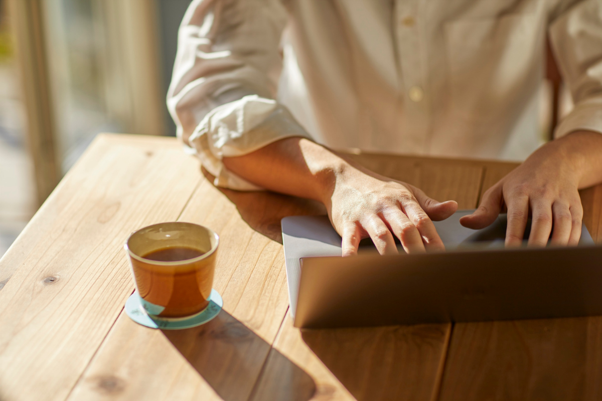 Person typing on a laptop at a sunlit wooden table with a cup of tea beside them.