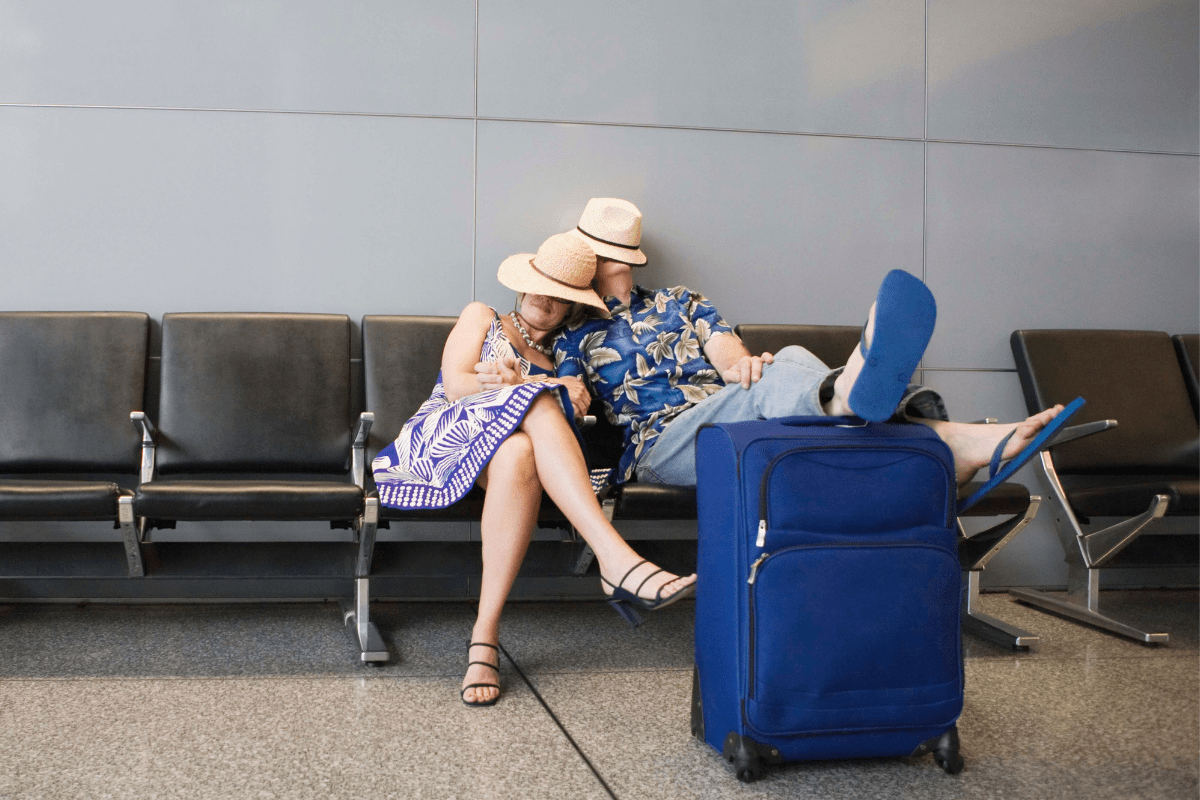 Couple sleeping in airport chairs with luggage.