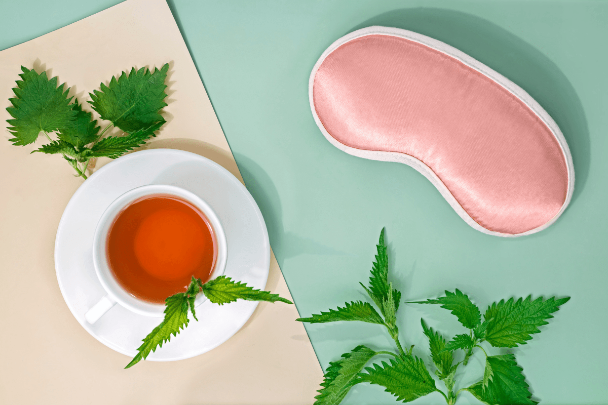 A cup of tea with fresh leaves placed beside a pink silk sleep mask on a pastel background.