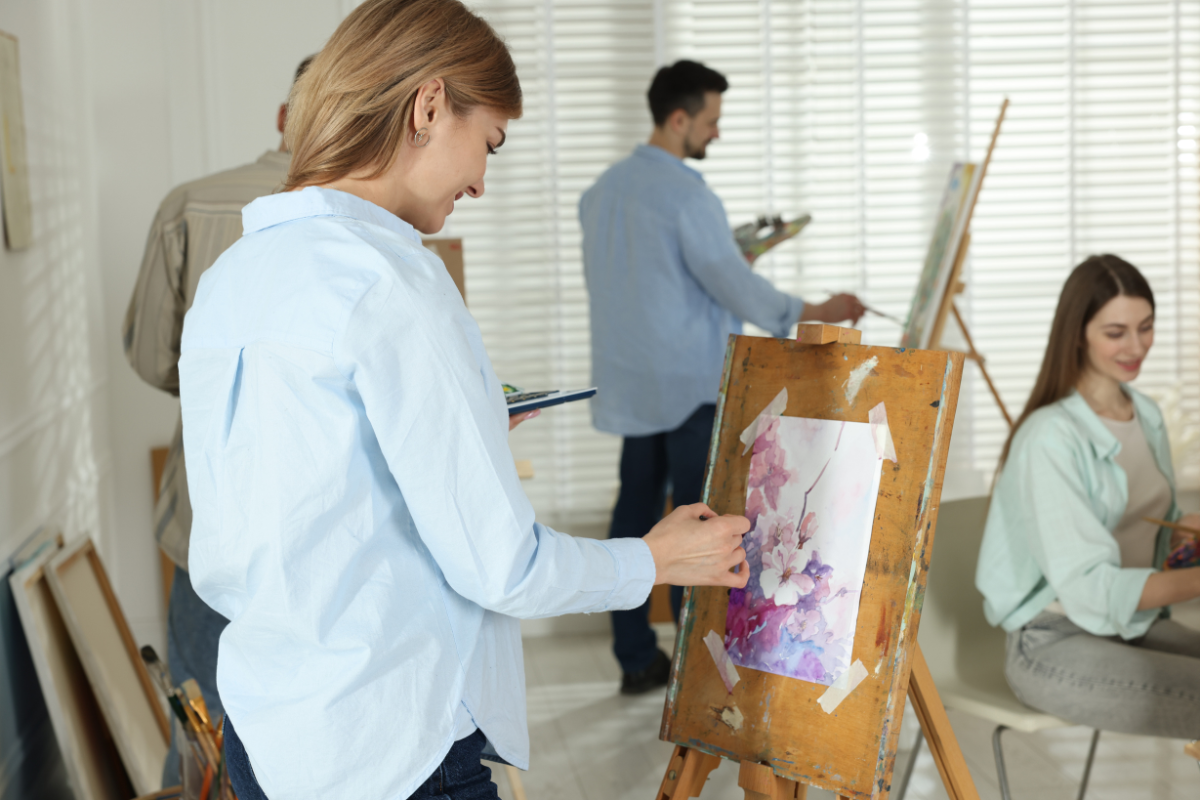 People painting at easels in a bright art class, one woman working on a watercolour of pink blossoms.