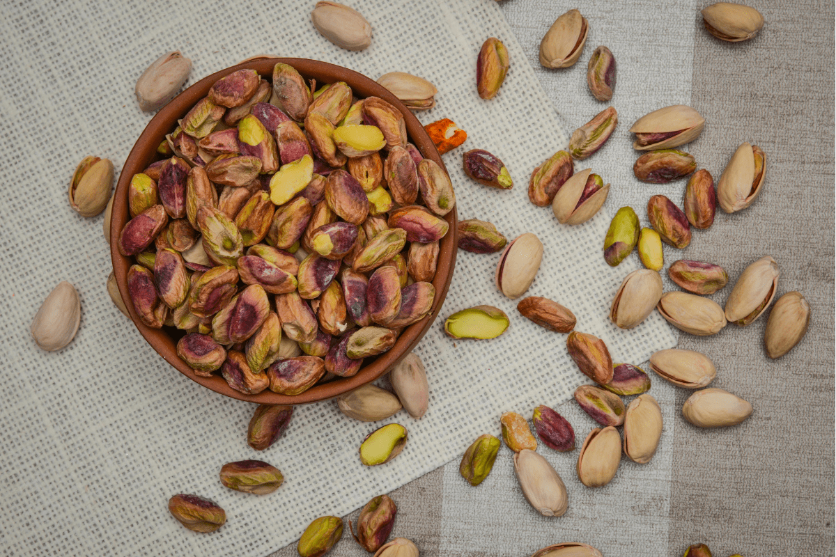 A bowl of shelled pistachios surrounded by whole pistachio nuts on a textured cloth.