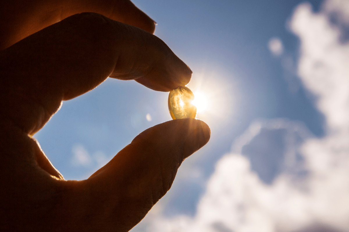 Hand holding a vitamin D capsule against sky with sunlight.