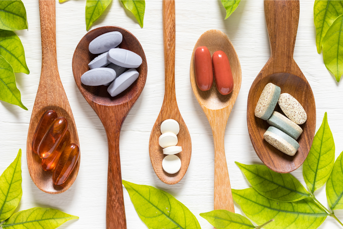 Various dietary supplement pills and capsules displayed on wooden spoons, surrounded by green leaves.