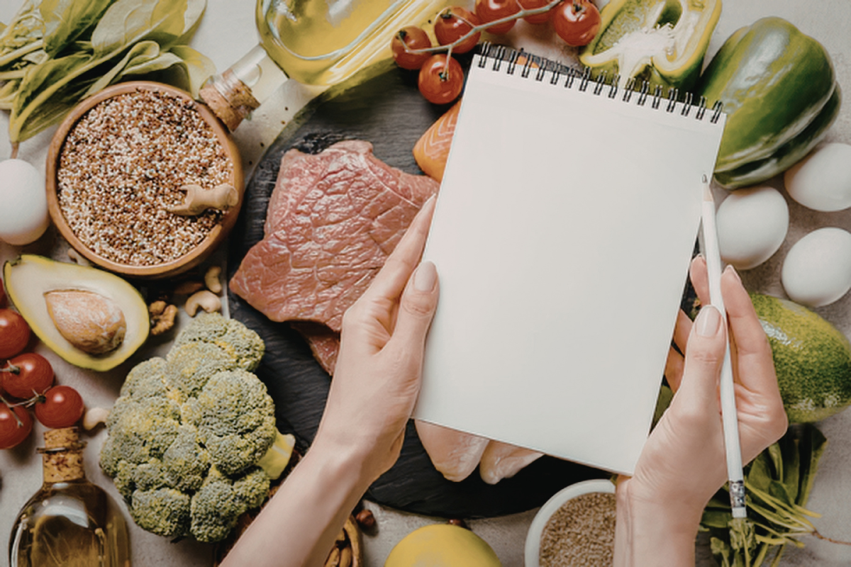 Hands holding a notebook over a spread of healthy foods including vegetables, meat, eggs, and grains.