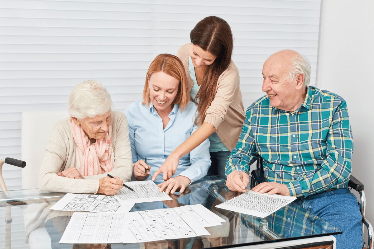 Two elderly people and two younger women solving puzzles together at a table.