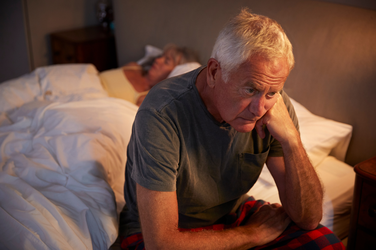 Older man sitting awake in bed while partner sleeps.