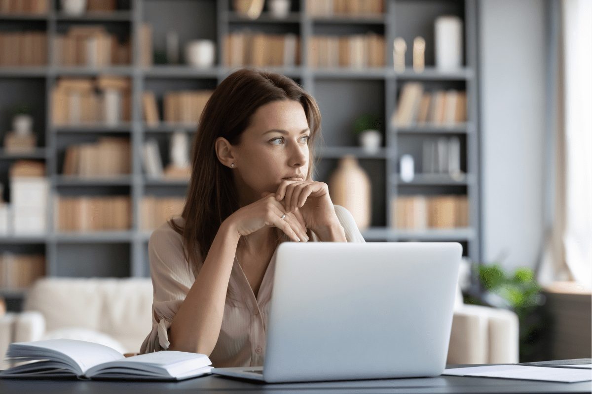 Woman staring thoughtfully while seated at a desk with a laptop.