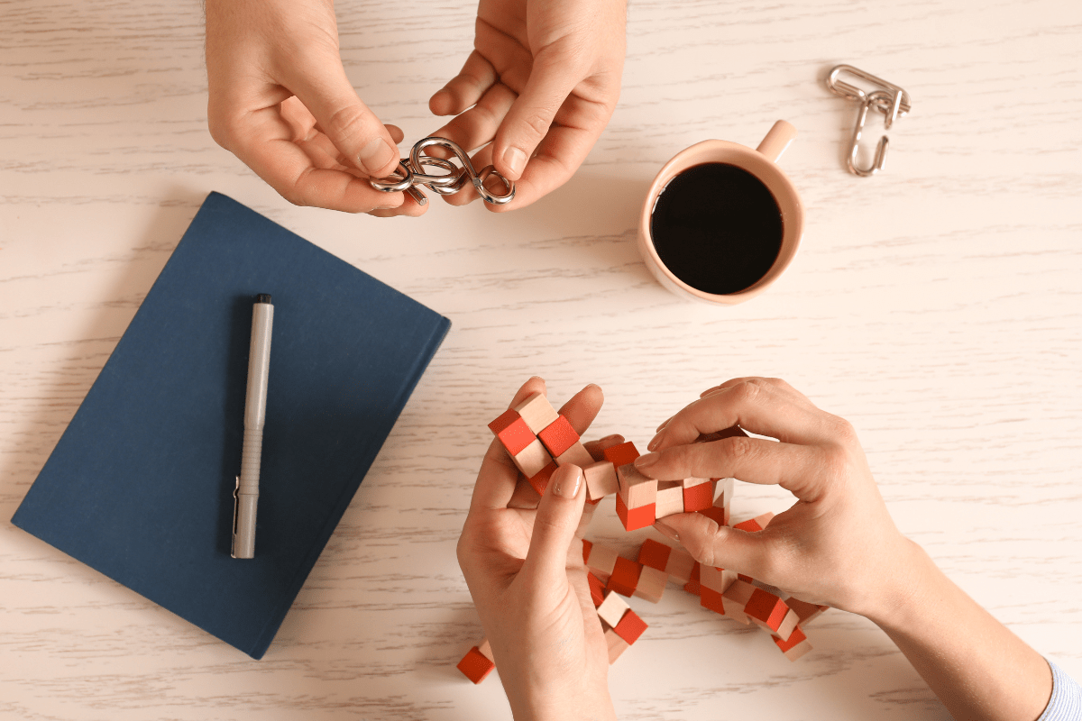 Hands solving puzzles and holding a coffee mug on a desk with a notebook and pen.