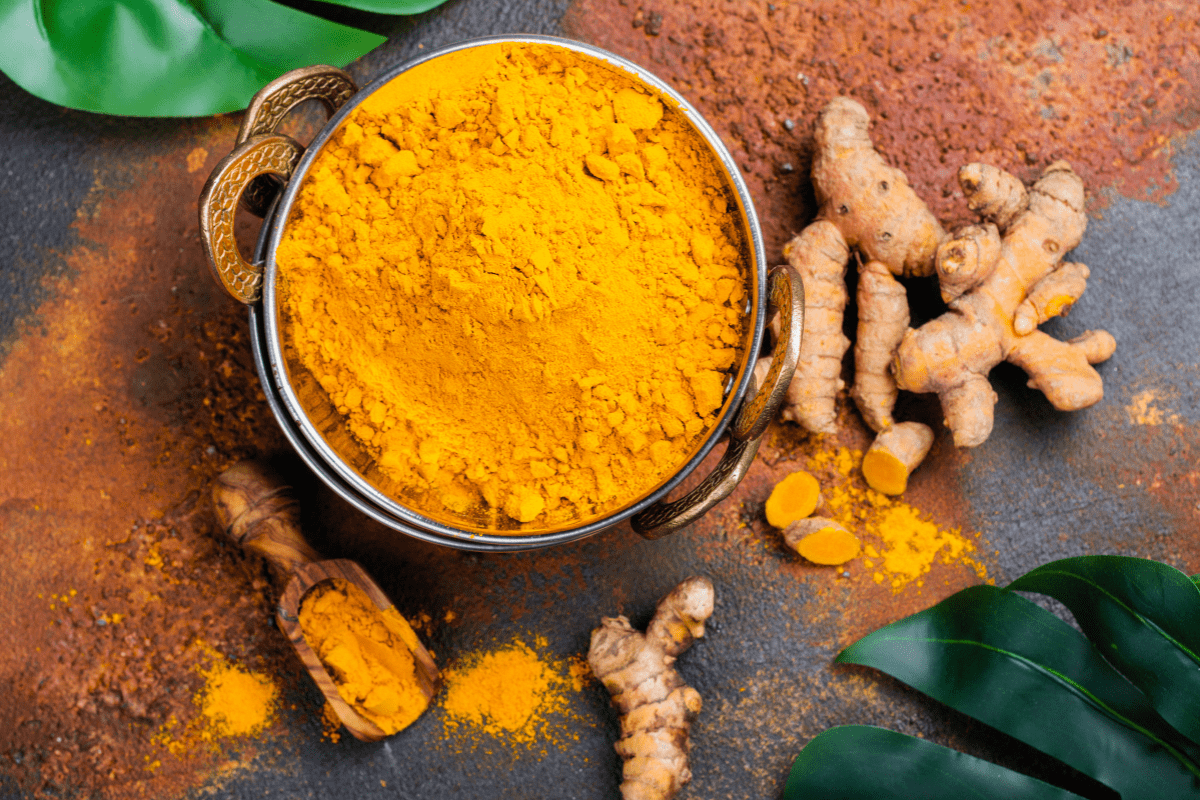 A bowl of turmeric powder with fresh turmeric roots on a textured surface.