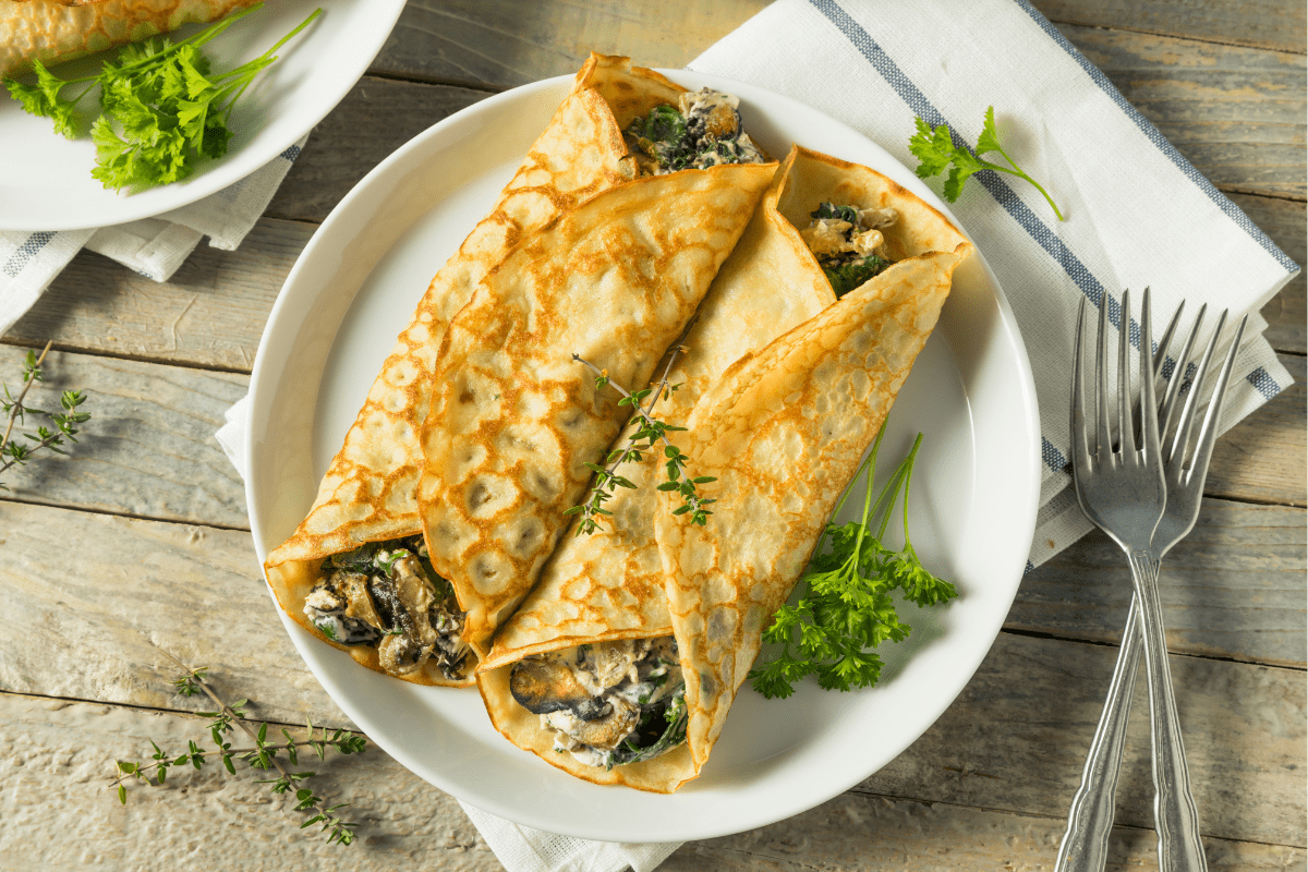Savoury crêpes filled with mushrooms and leeks, served with fresh herbs on a white plate.
